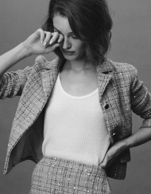 A young woman in a tweed jacket and skirt poses in a studio with her hand near her face, creating a classic, monochrome fashion portrait.