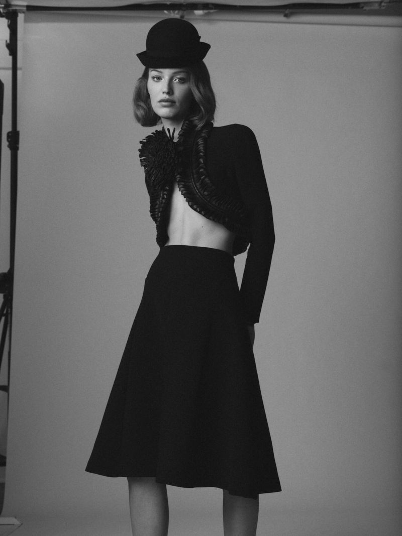 Model Maartje Convens in a black cropped jacket, black midi skirt, and bowler hat standing against a studio backdrop.