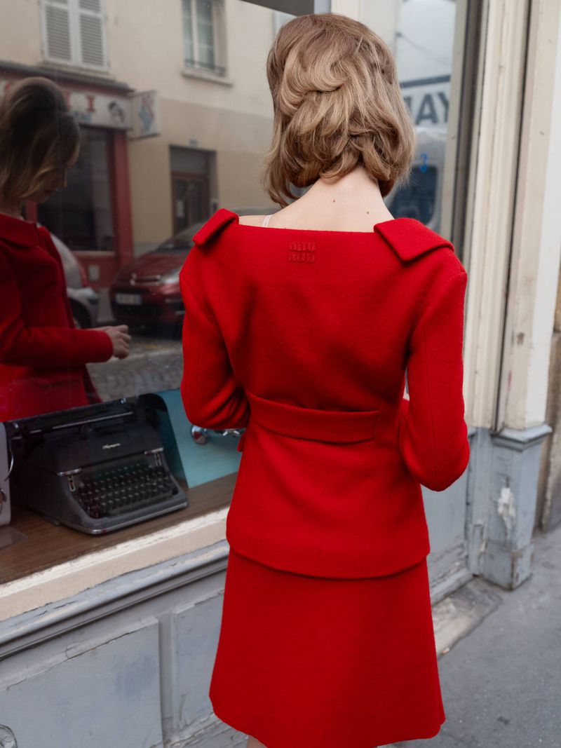 Back view of woman in a red skirt suit standing outside a building with reflective glass. A vintage typewriter is visible in reflection.