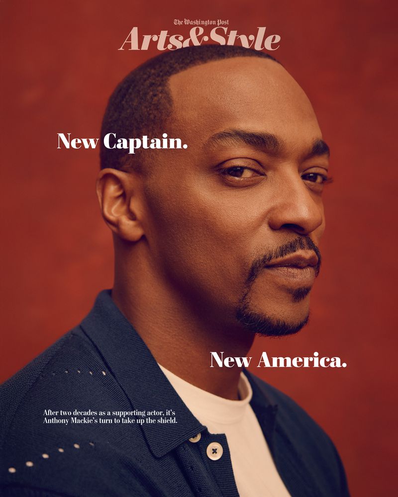 Close-up shot of Anthony Mackie, an adult male, wearing a dark blue shirt. The background is a solid reddish-brown color. The image is a portrait.