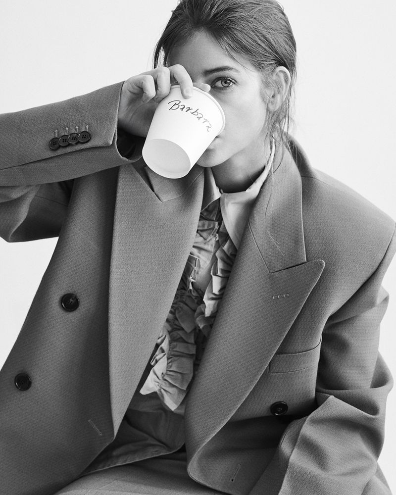 Monochrome portrait of a woman in an oversized blazer holding a paper cup with the name 'Barbara' on it, set against a white backdrop.