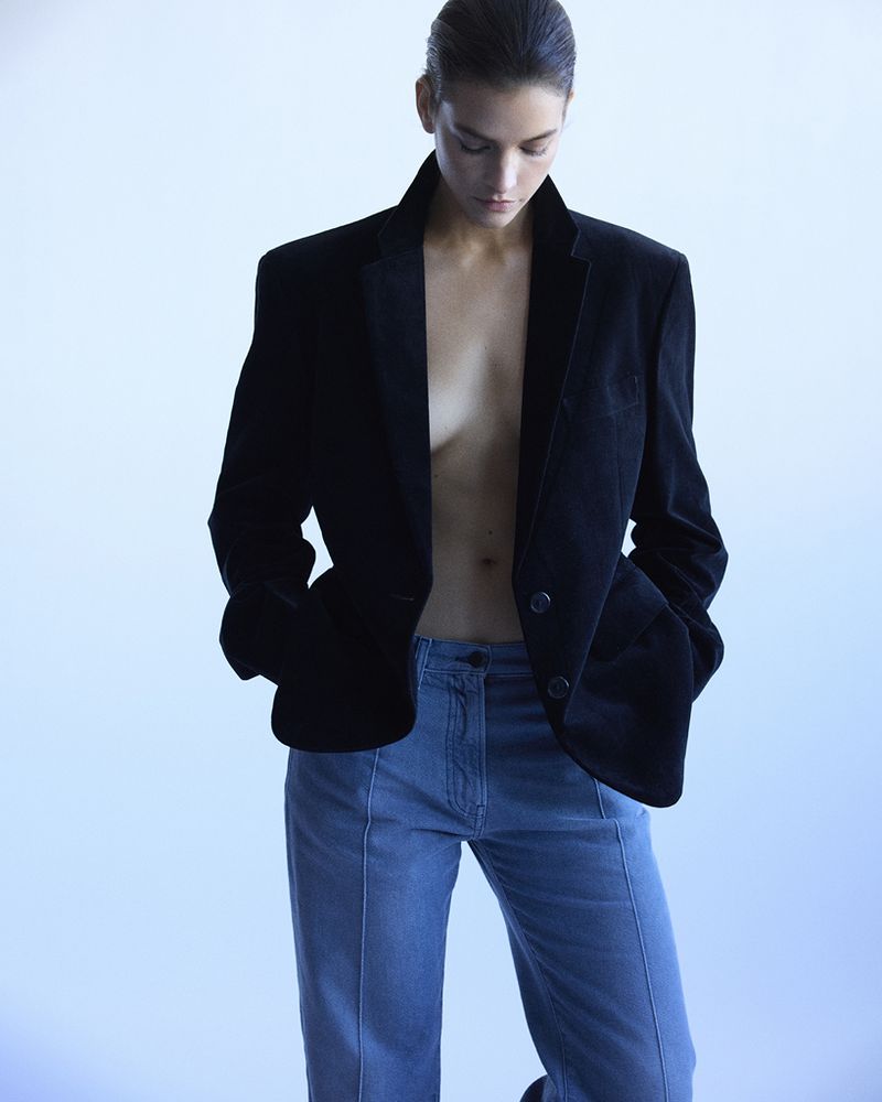 A woman posing indoors wearing a black velvet blazer and blue jeans looking down. The image features studio lighting and a light blue background.
