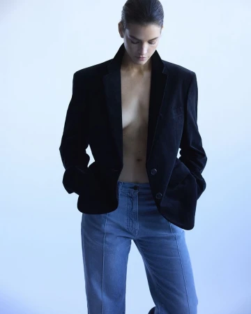 A woman posing indoors wearing a black velvet blazer and blue jeans looking down. The image features studio lighting and a light blue background.