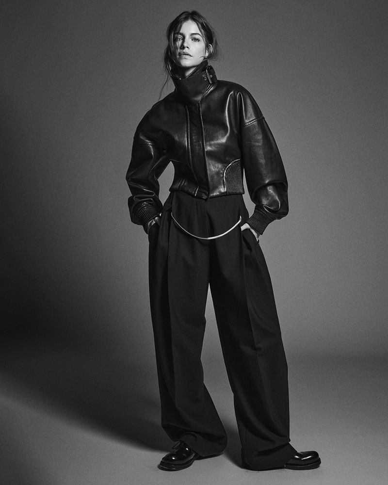 Black and white fashion shot of a woman in a leather jacket and wide-leg trousers in a studio, captured in a medium shot.