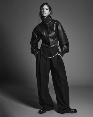 Black and white fashion shot of a woman in a leather jacket and wide-leg trousers in a studio, captured in a medium shot.