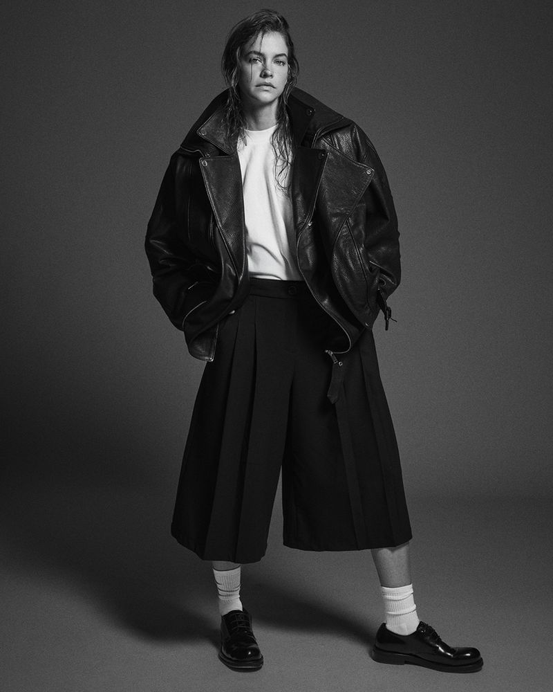 A woman in a black leather jacket, white tee, and black culottes strikes a pose in a studio with a neutral gray background.
