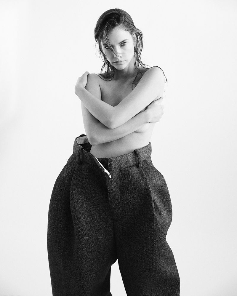 Black and white portrait of an adult woman with arms crossed, wearing oversized trousers, set against a bright background in a studio.