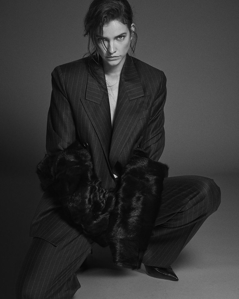 A young adult woman poses in a pinstripe suit with black fur cuffs in a studio setting with a serious expression, high fashion vibe.