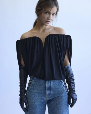 Barbara Palvin wearing an off-the-shoulder black top and leather gloves with denim jeans against a plain white wall