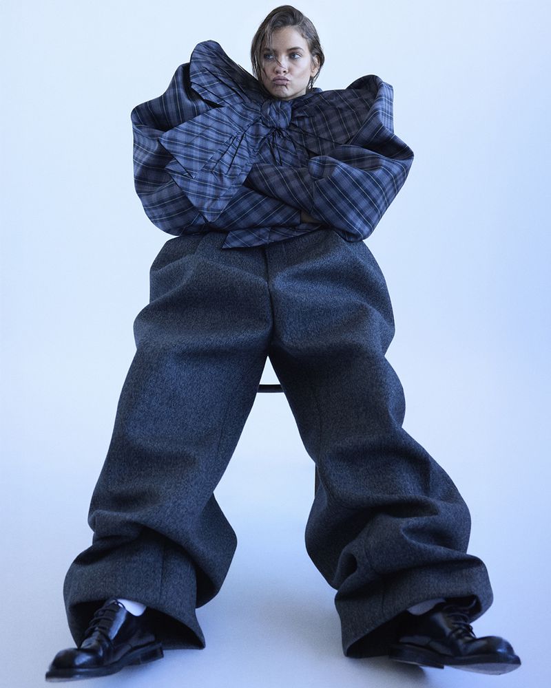 A young adult woman with a neutral facial expression sits in an unusual oversized grey plaid top and baggy grey pants, and black shoes.
