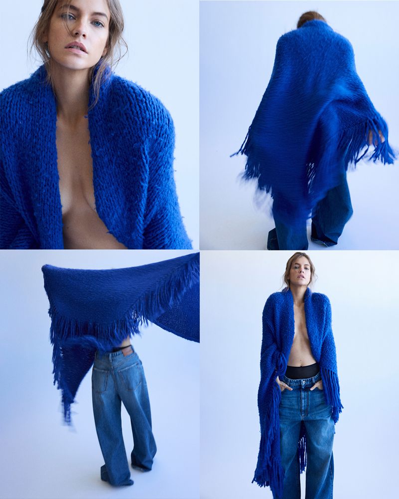 A woman in a stylish blue knit shawl and jeans poses in a photo studio. The fashion shot has a bright, clean aesthetic and soft lighting.