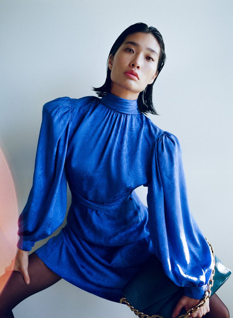 Asian woman models a blue mini dress with long sleeves, paired with dark tights and a shiny green handbag with gold chain.