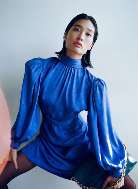 Asian woman models a blue mini dress with long sleeves, paired with dark tights and a shiny green handbag with gold chain.