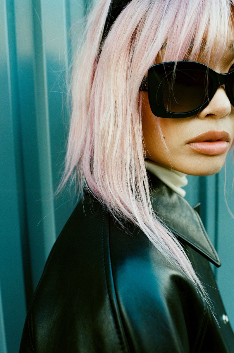 Close-up of a woman wearing a black leather jacket, black sunglasses, and a white turtleneck sweater with pink hair and a black headband.