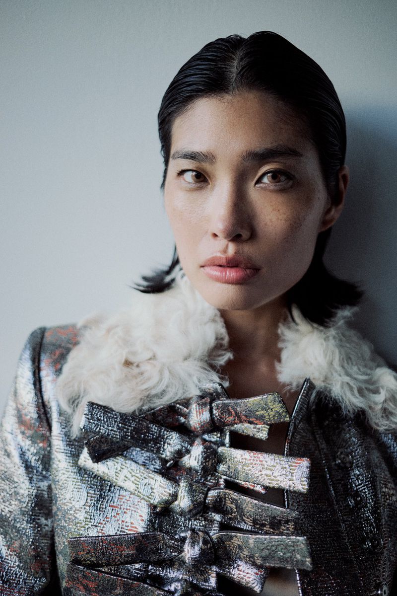 Close-up studio shot of an Asian woman wearing a metallic silver jacket with a fur collar and bow details, with slicked-back hair.