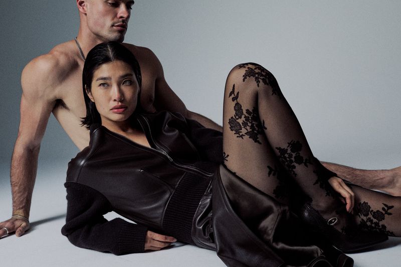 A woman in a jacket and floral patterned tights reclines with a bare-chested man against a neutral backdrop, studio lighting, fashion shot.