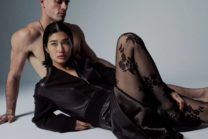 A woman in a jacket and floral patterned tights reclines with a bare-chested man against a neutral backdrop, studio lighting, fashion shot.