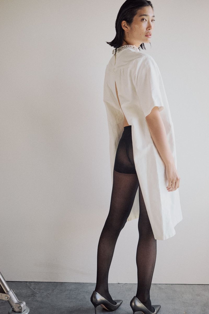 A fashion shot of an Asian woman in a cream dress, black tights, and silver heels posing against a white backdrop, showcasing contemporary style.