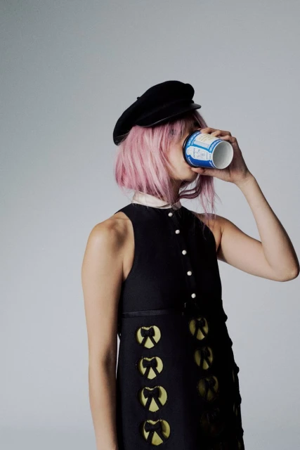 Stylish woman with pink hair wearing a black dress and hat, drinking from a paper cup against a grey background in a studio setting.