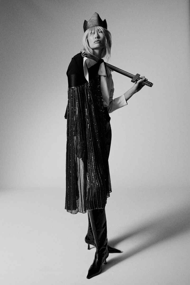 Fashion model poses in a studio wearing a cape with tassels, high boots, a unique hat, and holding a decorative sword in a studio setting.