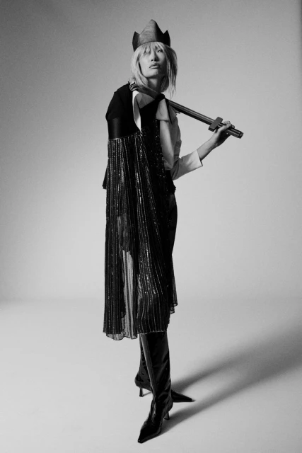Fashion model poses in a studio wearing a cape with tassels, high boots, a unique hat, and holding a decorative sword in a studio setting.