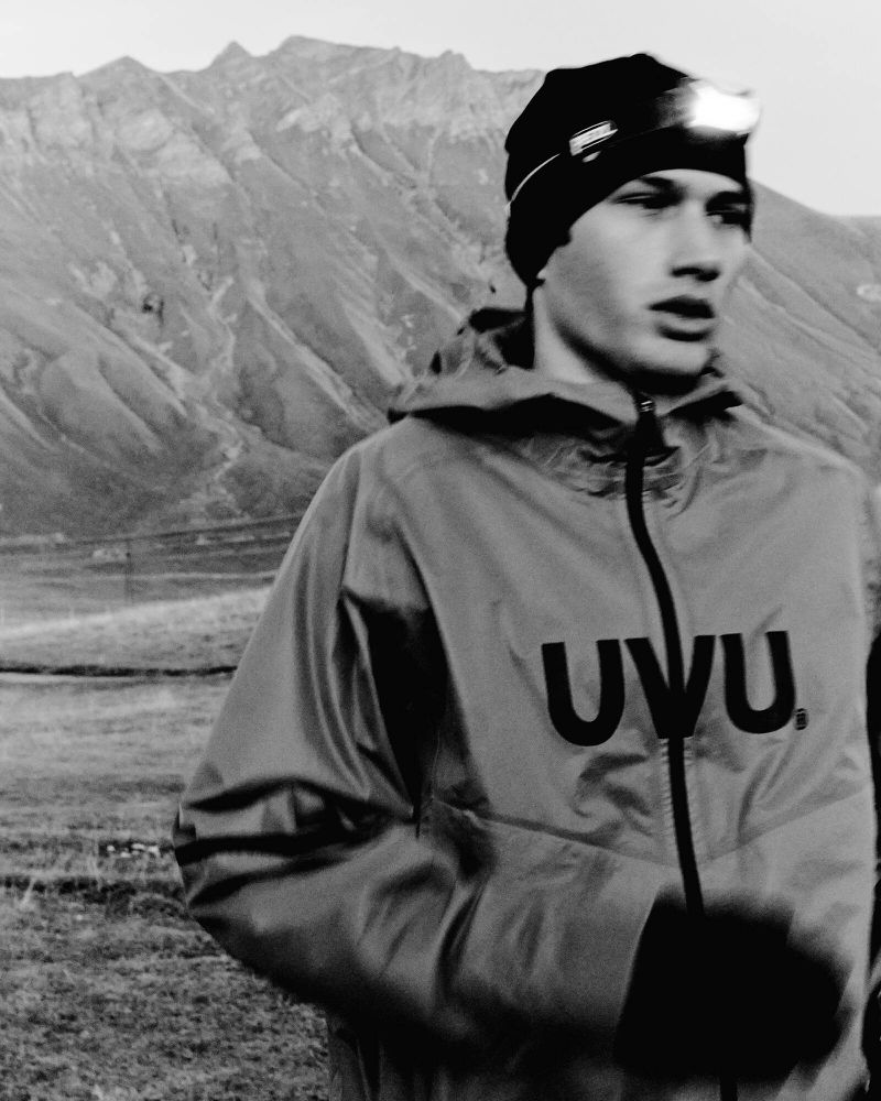 A young adult wearing a jacket and headlamp stands in a grassy field with rocky mountains in the background, creating a monochrome scene.