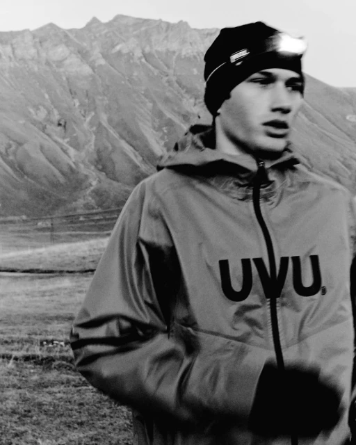 A young adult wearing a jacket and headlamp stands in a grassy field with rocky mountains in the background, creating a monochrome scene.