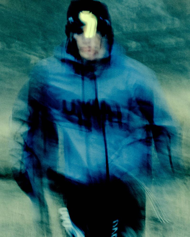 Abstract fashion portrait. A figure in motion wears a blue jacket with black text on the front and a light over the eyes.