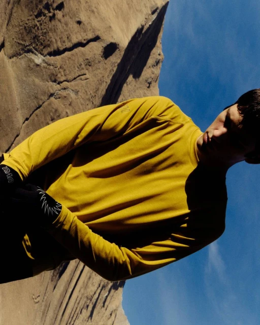 Man in yellow shirt poses in front of rock cliff and sky. Low angle shot. Outdoor setting with bright sunny day, fashion and fitness. 