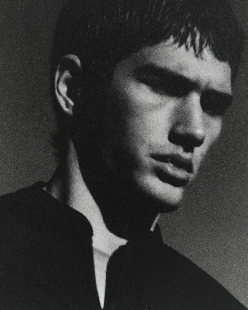 Monochromatic portrait of a young adult male with dark hair, wearing a dark collared shirt, gazing downward in a photo studio setting.