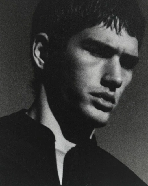 Monochromatic portrait of a young adult male with dark hair, wearing a dark collared shirt, gazing downward in a photo studio setting.