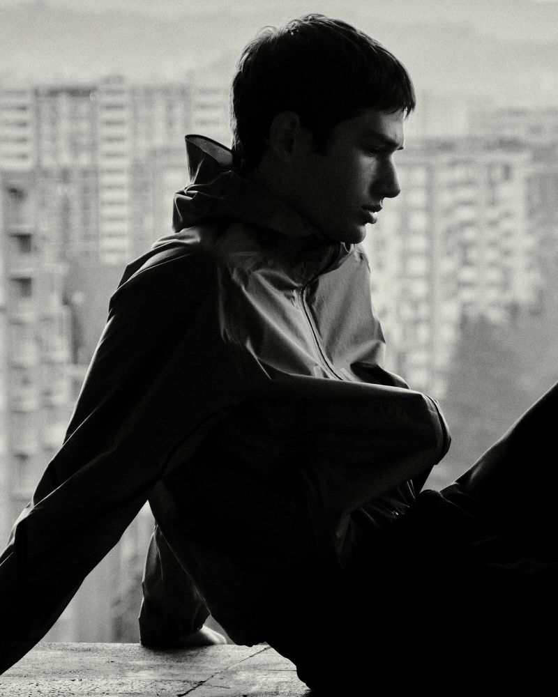 Monochrome portrait of a young man wearing a zipped jacket against a blurred cityscape background.