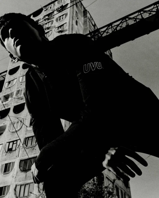 Low-angle shot of a man in a black sweatshirt with 'UVU' on the sleeve, with a tall building and metal structure in the background against a gray sky.