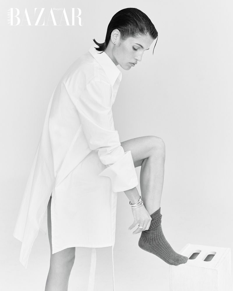 Black and white studio portrait of a woman in an oversized white shirt putting on a sock, set against a plain backdrop, fashion editorial.