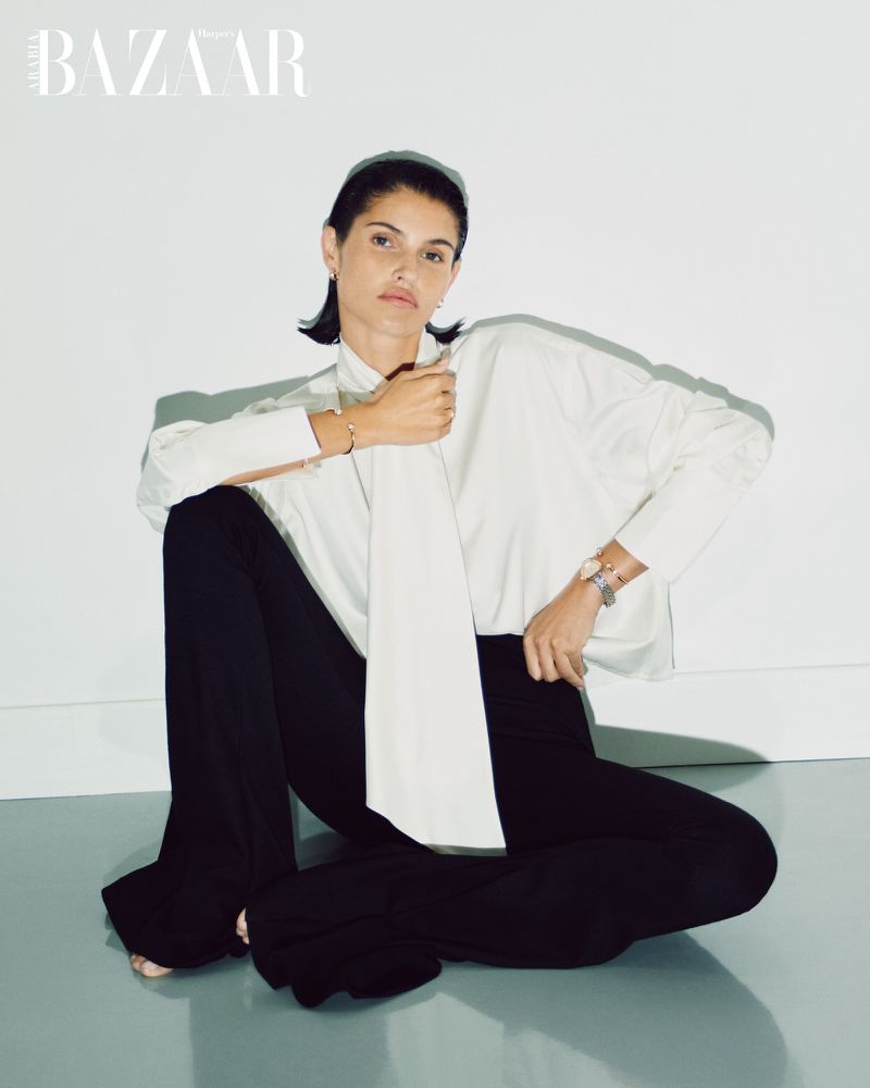 Elegant woman poses indoors wearing a white, collared shirt and black pants. She sits on a gray surface against a white background.