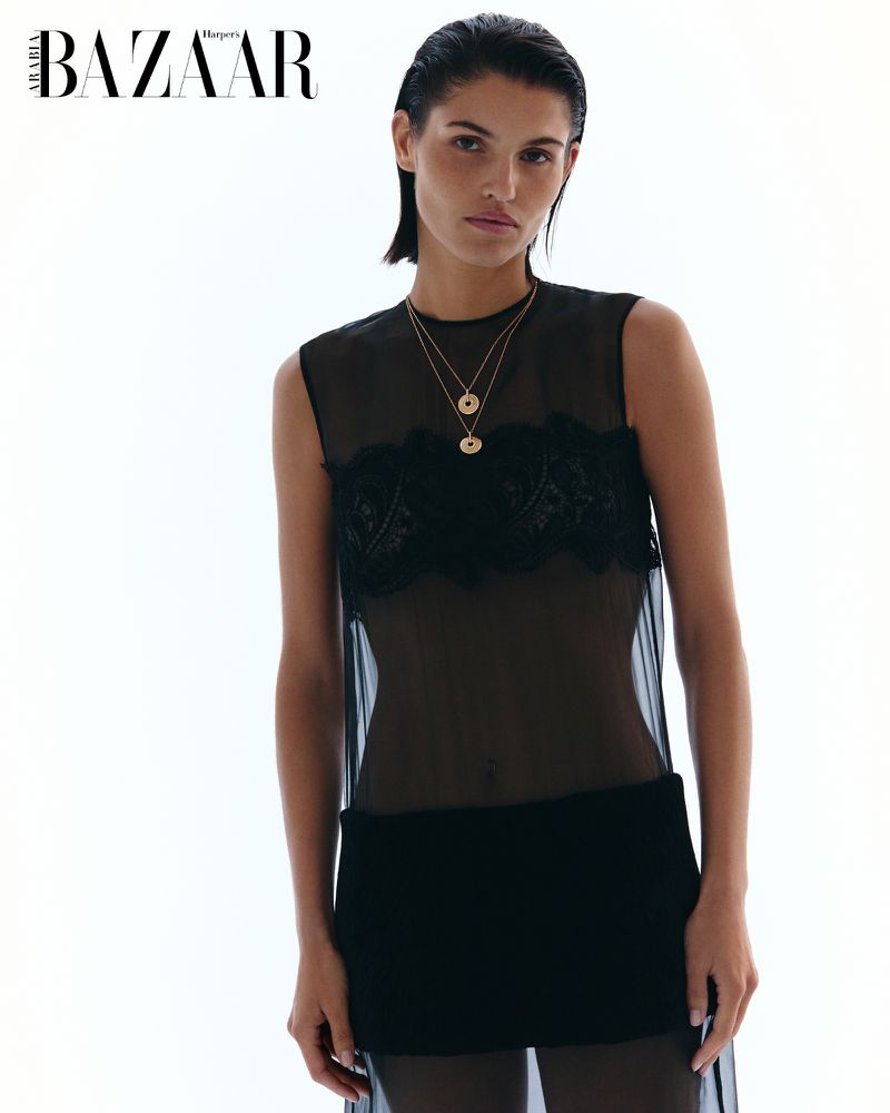 A woman poses in a black dress in front of a bright white background for Harpers Bazaar. She wears two gold pendant necklaces.