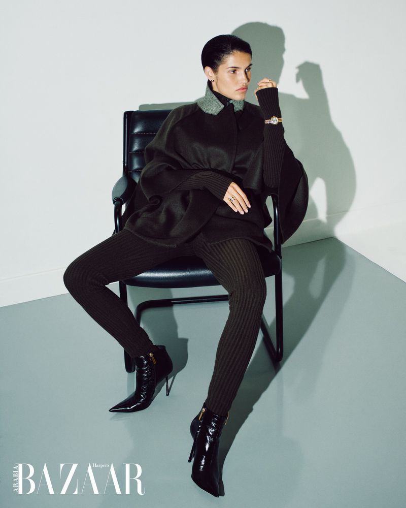 Fashion editorial shot of a woman in a dark brown outfit and black boots sitting in a black chair in front of a light gray wall.
