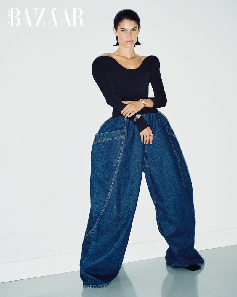 Fashion-forward woman in black top and oversized blue denim pants poses against a white wall, showcasing a unique style and silhouette.