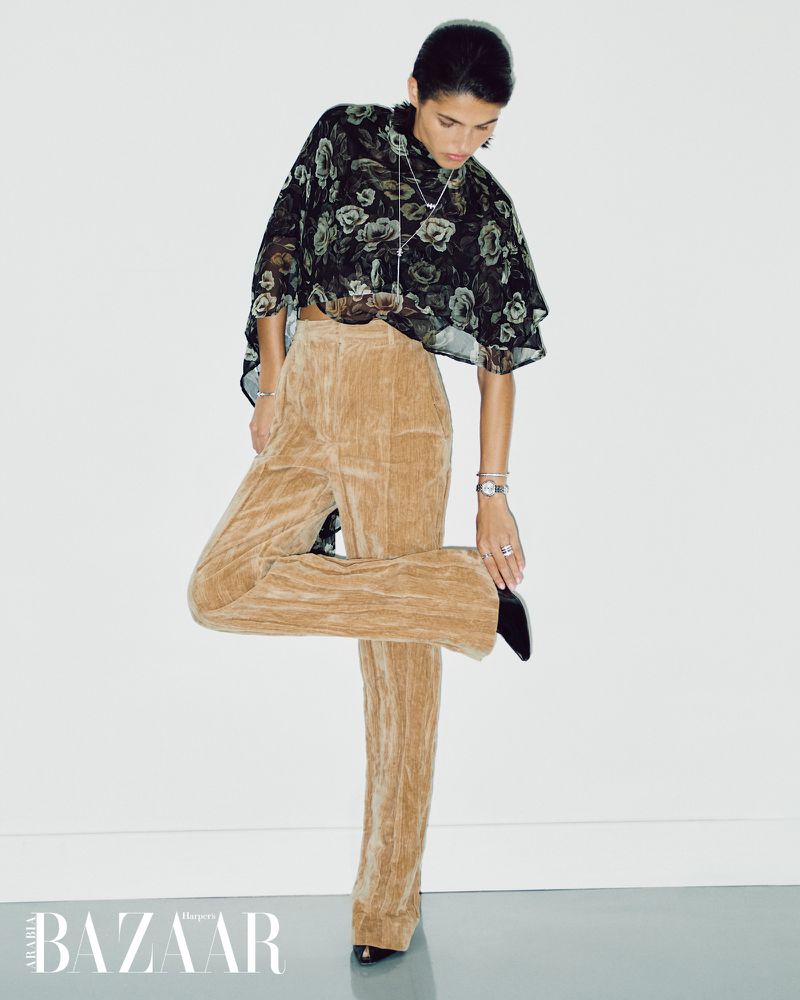 Fashion shot of a woman in a floral blouse and beige pants, posing in front of a white wall with a magazine logo at the bottom.