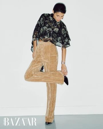 Fashion shot of a woman in a floral blouse and beige pants, posing in front of a white wall with a magazine logo at the bottom.