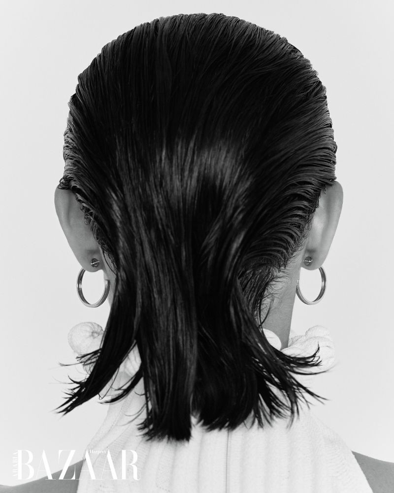 Back view of a woman with slicked-back, dark wet hair, wearing hoop earrings and a white top, against a plain white background.