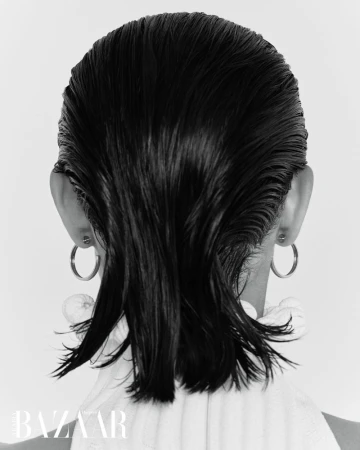 Back view of a woman with slicked-back, dark wet hair, wearing hoop earrings and a white top, against a plain white background.