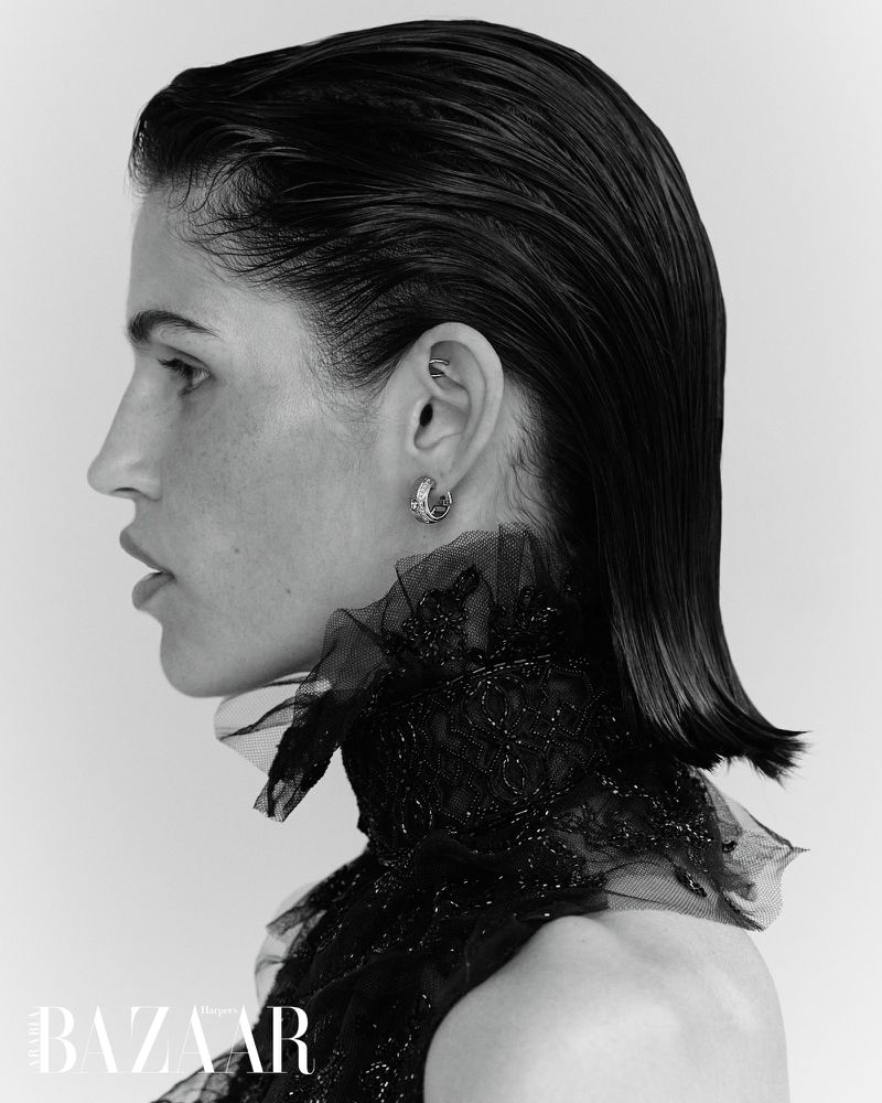 Monochromatic portrait of a woman with wet-look hair, wearing an elegant black lace high-neck dress, earring, and a neutral background.