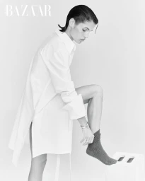 Model wearing an oversized white shirt pulling up a sock with one foot on a white box against a seamless background.