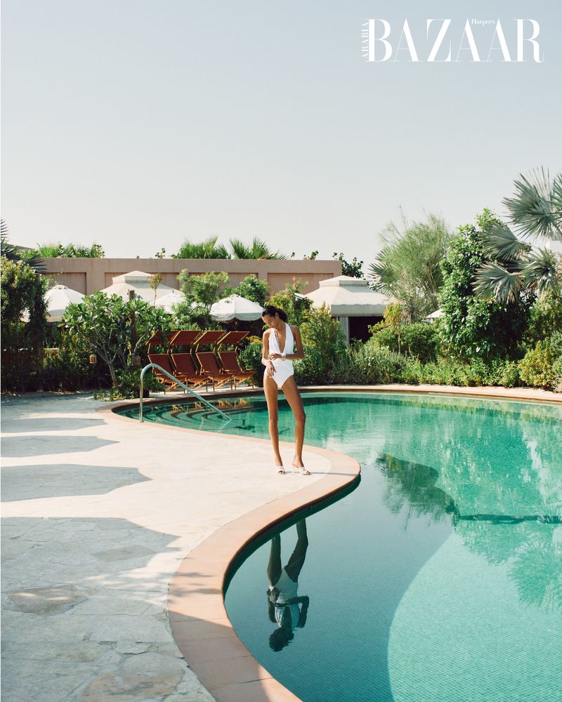 Woman in a white bathing suit stands by a turquoise pool surrounded by lush greenery on a sunny day, Harpers Bazaar logo visible.