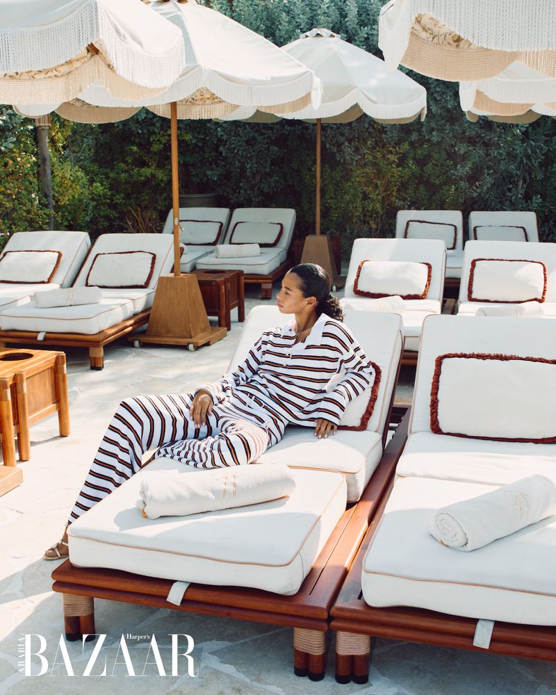 Woman lounges on a wooden sunbed under fringed parasols at an outdoor resort, wearing a striped pajama set on a sunny day.