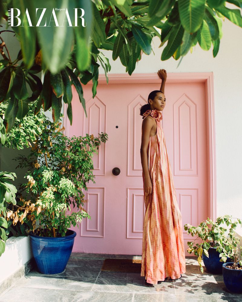 Elegant adult female fashion model poses in a long patterned dress, next to a pink door with plants under a green leafy tree canopy.