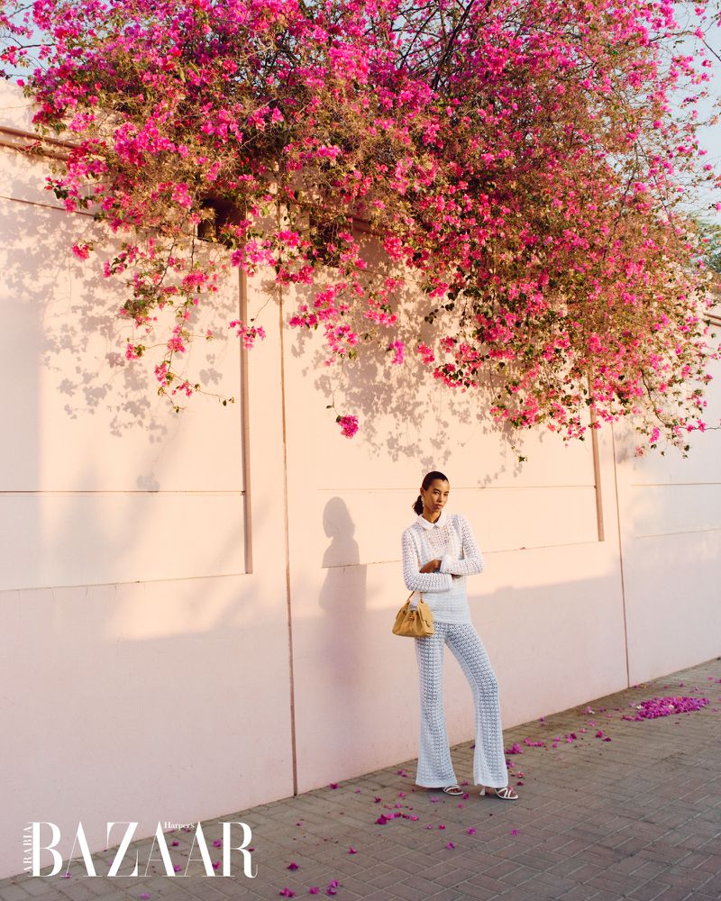 Fashion editorial shot of a woman posing against a wall with flowering vines, wearing a white knit outfit and yellow purse, outdoors.