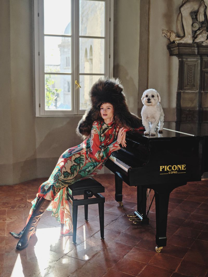 Woman in patterned dress and fur hat poses next to grand piano with small white dog in room with tile floor and window.