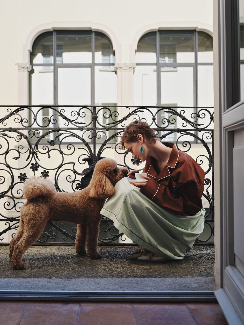 Woman in pajamas kneels with a brown poodle on a balcony with wrought iron railing and a cream facade building in the background.